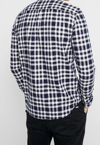 Camicia a scacchi blu navy e bianco, maniche lunghe, taglio dritto con colletto a bottoni e tasca laterale, realizzata in tessuto leggero.