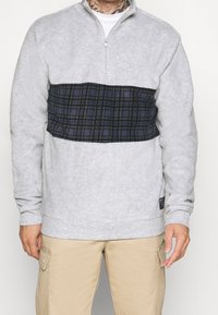 Grå fleece-tröja med halvdragkedja, med en marinblå rutig panel över bröstet. Strukturvävt tyg och ribbade manschetter.