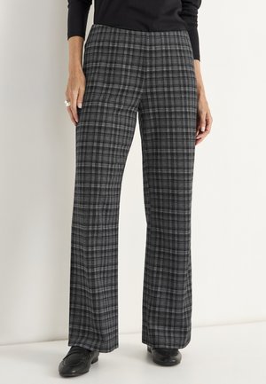Vrouw in grijs en zwart geruite pantalon met rechte benen en zwarte instappers, staand tegen een effen lichte achtergrond.