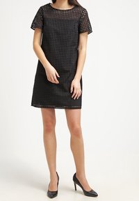 Vestido negro con un diseño de celosía abierta, mangas cortas y una silueta recta. Combinado con zapatos de tacón altos negros.