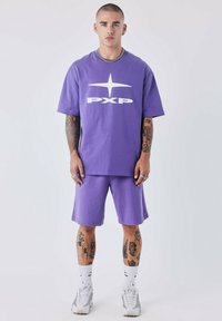 Paarse t-shirt met groot wit "PXP" logo, gecombineerd met bijpassende paarse shorts. Katoenen stof, relaxed fit, met een zilveren ketting als accent.