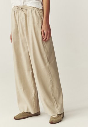 Osoba nosiaca béžové široké nohavice s elastickým pásom a béžové tenisky, stojaca pred jednoliatym svetlým pozadím.