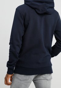 Sudadera con capucha de color azul marino, hecha de una tela suave, presenta un bolsillo canguro y puños acanalados, mostrada desde la parte posterior con un diseño minimalista.