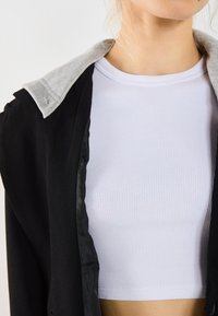 Čierna bunda s šedým golierom, nosená cez biely rebrovaný crop top. Bunda má hladkú textúru a mierne voľný strih.