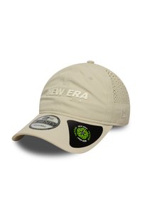 Beige New Era sapka, amely könnyű anyagból készült, oldalsó szellőzőnyílásokkal, domború logóval és állítható szalaggal rendelkezik. Zöld és fekete címkék csatlakoznak hozzá.