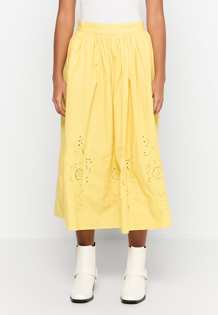 Falda midi de algodón amarilla con cintura fruncida, que presenta detalles de bordado en ojales. Llevada con botines blancos. Textura suave.