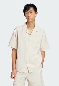 Short-sleeve cream button-up shirt with a patch pocket, textured fabric, and subtle logo detail on the pocket. Coordinating lightweight shorts.