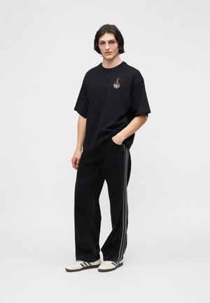 Άνδρας που φοράει μαύρο φαρδύ T-shirt με μικρό ροζ σχέδιο, χαλαρό μαύρο παντελόνι με πλαϊνές ρίγες, λευκά αθλητικά παπούτσια και ροζ γυαλιά στέκεται όρθιος.