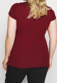 Kvinna med blont hår iförd en kortärmad, ribbad vinröd topp och svarta byxor, fotograferad bakifrån mot en slätt bakgrund.