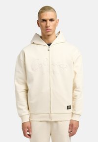 Creme zip-up hættetrøje lavet af blødt stof, med broderet tekst foran, snøre i hætten og ribbede manchetter og kant.