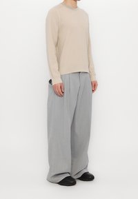 Beżowy sweter dziany z okrągłym dekoltem, zestawiony z szerokimi szarymi spodniami z plisami oraz czarnymi butami. Gładka faktura i luźny krój.