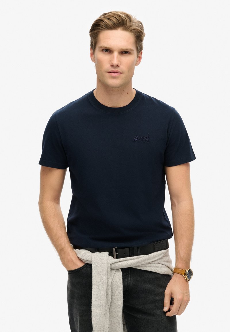 Mand med lysebrunt hår iført en marineblå T-shirt, sorte jeans og en grå sweater bundet om livet, stående med hænderne i lommerne.