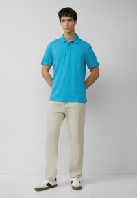 Turquoise poloshirt met een klassieke kraag en opgerolde mouwen, gecombineerd met lichtbeige broek en sneakers met zwarte accenten.