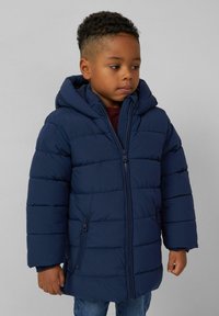 Jong kind met korte krullende haren, gekleed in een navyblauwe, ritssluitende, gevoerde jas met capuchon en blauwe spijkerbroek, kijkt opzij.