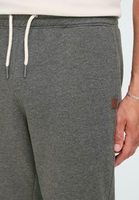 Grå sweatpants med myk tekstur, som har snøring i kremfarge og en liten brun logo-lapp på venstre side.