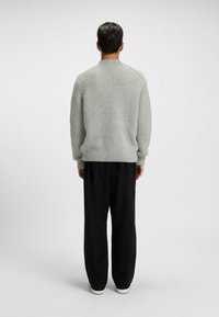 Hellgrauer Strickpullover mit einem strukturierten Muster, gerippten Bündchen und Rundhalsausschnitt, kombiniert mit lockeren schwarzen Hosen und weißen Sneakers.
