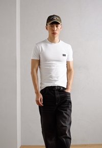 T-shirt bianca a maniche corte con un piccolo etichetta nera, abbinata a jeans scuri e un cappellino verde. Il cappellino presenta un logo in colori chiari.