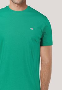 Grünes Baumwoll-T-Shirt mit kurzen Ärmeln, rundem Halsauschnitt und kleinem gesticktem Logo auf der Brust. Glatte Textur und taillierte Passform.