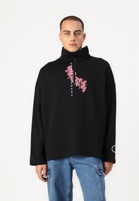 Black zip-up sweatshirt with a high collar, featuring pink floral graphics and white text. Loose fit. Paired with blue denim pants.