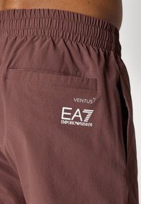 Smeđe kratke hlače s elastičnom sredinom struka, s stražnjim džepom i bijelim logotipom "EA7 EMPORIO ARMANI" otisnutim blizu struka. Glatka tekstura.