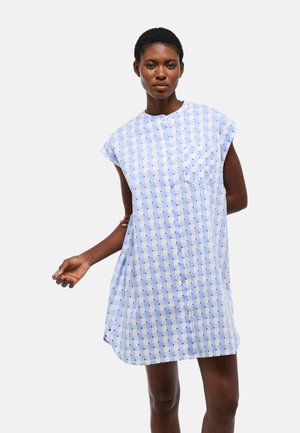 Femme portant une robe boutonnée à manches courtes avec un motif à carreaux bleu et blanc et de petits pois rouges, debout devant un fond clair uni.