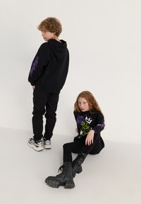 Sweatshirt preto com capuz e texto roxo, combinado com calças pretas e ténis chunky. O segundo conjunto apresenta uma t-shirt gráfica preta e botas de combate.