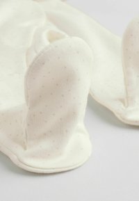 Cremefarbene Stoffschühchen mit weicher Textur, versehen mit kleinen grauen Tupfen und abgerundeten, plüschigen Ohren oben.