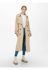 Beige trenchcoat med store sorte knapper, brede klipper og sidelommer, båret over en hvid bluse og blå jeans, sammen med cremede støvler.