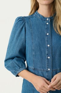 Denim dress in medium blue with a high collar, puff sleeves, and button closures down the front. Features a relaxed fit and stitch detailing.