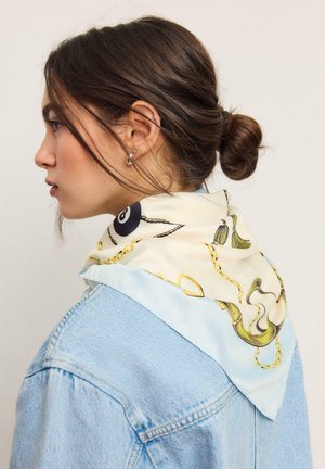 Žena smeđom kosom u punđi nosi svijetloplavu traper jaknu i svijetli svileni šal s uzorcima lanaca i resica oko vrata.