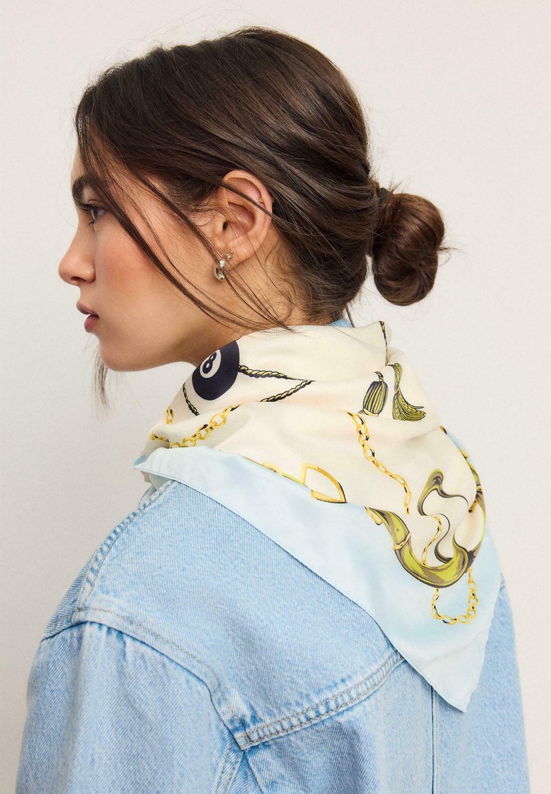 Femme aux cheveux bruns attachés en chignon, portant une veste en jean bleu clair et un foulard en soie de couleur claire avec des motifs de chaînes et de glands autour du cou.