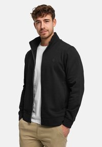 Svart jacka med dragkedja, gjord av slät tyg, med hög krage, ribbade ärmslut och en diskret logotyp på bröstet. Bärs över en vit t-shirt.