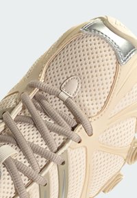 Svetlo béžová teniska s hornou sieťovinou, zaokrúhlenou špičkou, plochými šnúrkami, kovovými zdobeniami a gumovou podrážkou s charakteristickými dizajnovými prvkami.