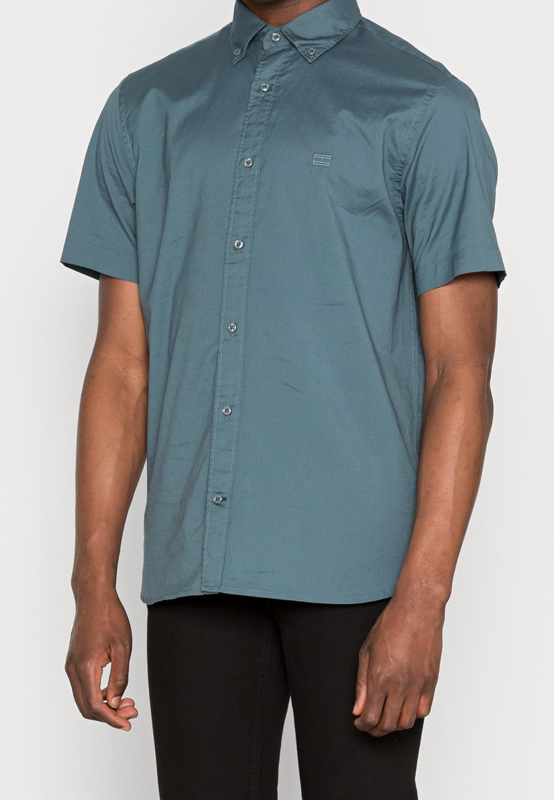 Camicia a maniche corte, con bottoni, in tessuto teal con un leggero lucido. Caratterizzata da un piccolo logo, una texture morbida e un design del colletto classico.
