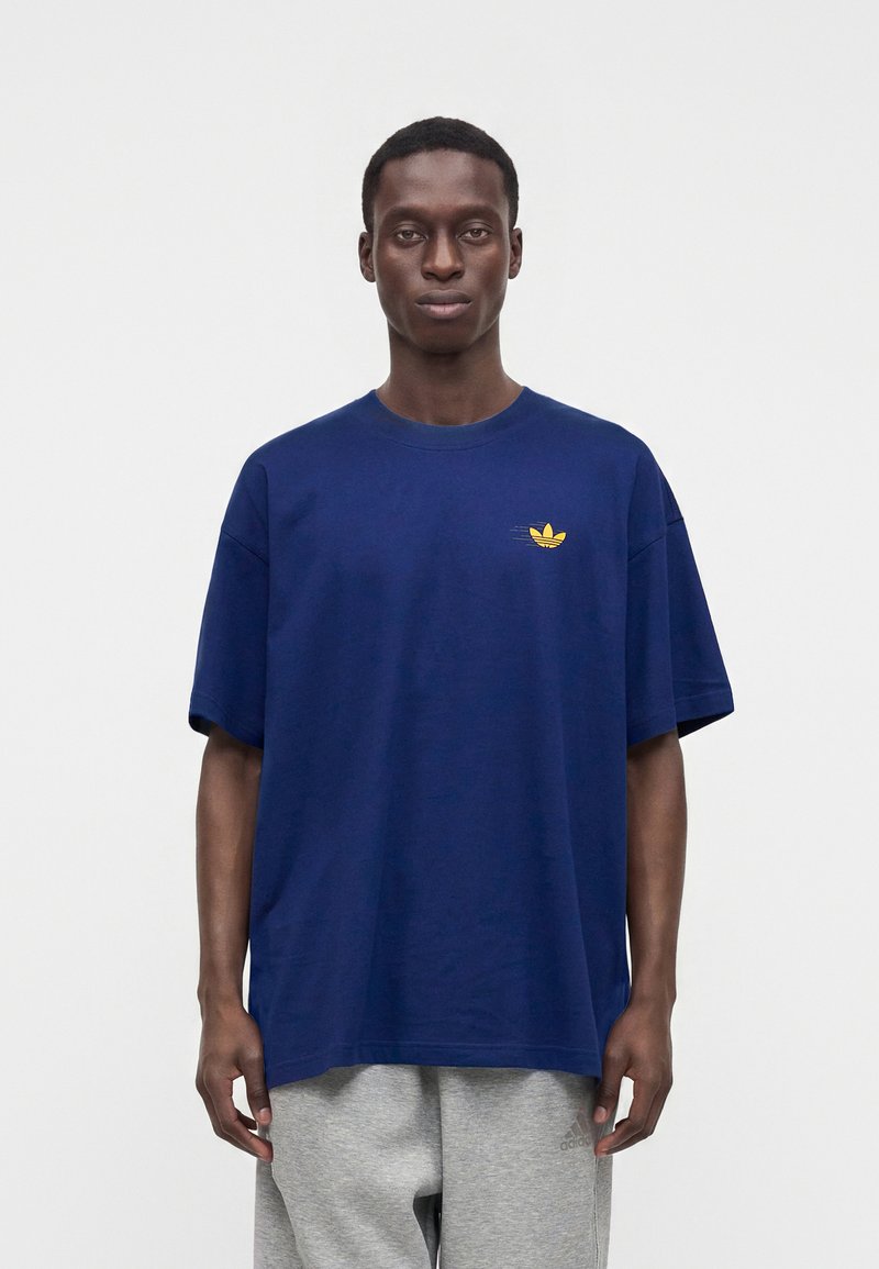 Hombre de pie con una camiseta suelta de color azul oscuro con un pequeño logo amarillo en el pecho y pantalones deportivos gris claro.