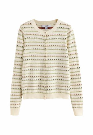Junge Frau trägt eine cremefarbene, gemusterte Strickjacke, weite hellolivfarbene Hosen und schwarze Ballerinas, steht auf einem Holzboden vor einer beigen Wand.