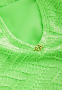 Neon-grünes Kleidungsstück mit Spitzenstruktur und geometrischem Muster sowie einem kleinen goldenen Verschlussdetail im Bereich des Ausschnitts.