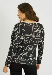 Haut à manches longues en tissu noir présentant un motif abstrait en lignes blanches de visages. Texture lisse avec un col rond et une coupe ample.