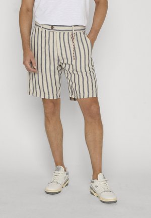 Man draagt crème- en marineblauwe verticale gestreepte korte broek met een gevlochten sleutelhanger, witte sneakers en een wit T-shirt, staand tegen een effen achtergrond.