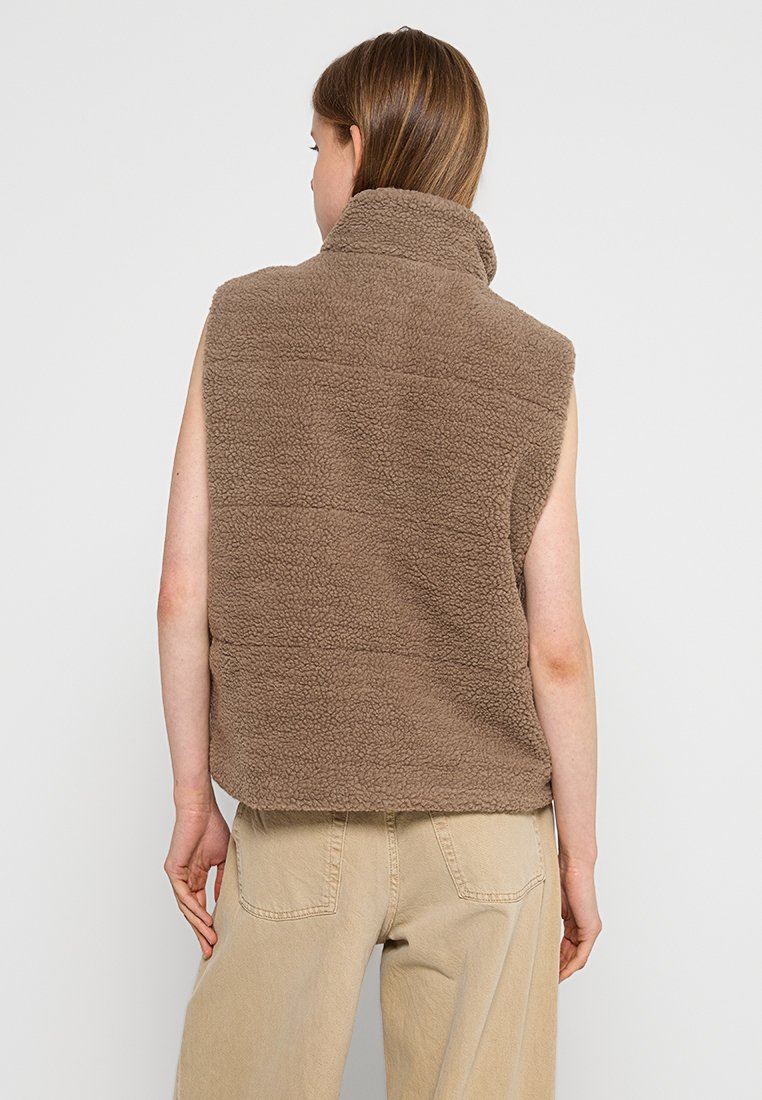 Gilet sans manches brun en polaire avec un col haut, surface texturée douce et détails de couture horizontaux. Associé à un pantalon beige clair.