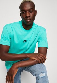 Tirkizna pamučna majica kratkih rukava s okruglim izrezom i crnim Nike logom na prsima, u kombinaciji sa svijetloplavim poderanim trapericama.