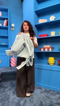 Ženska z kodrastimi lasmi drži svetlomodro jakno, stoji v modri sobi z policami, na katerih so knjige, ura in dekorativni predmeti.