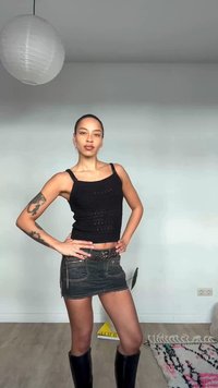 Noor tätoveeritud kätega, musta toppi, musta teksase miniseeliku ja mustade saabastega isik, kes poseerib enesekindlalt siseruumides.