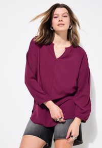 Bordeauxfarbige Bluse mit V-Ausschnitt, langen Ärmeln und strukturierter Stoff, kombiniert mit dunkelgrauen Denimshorts und silbernem Zubehör.