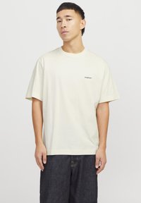 Kremfarget bomull t-skjorte med avslappet passform, korte ermer og en liten svart "Originals"-logo på brystet. Kledd med mørke denimshorts.