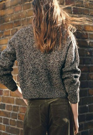 Suéter de punto marrón texturizado con un diseño recortado, combinado con pantalones de pana oliva que cuentan con bolsillos traseros, todo ello frente a un fondo de ladrillo.