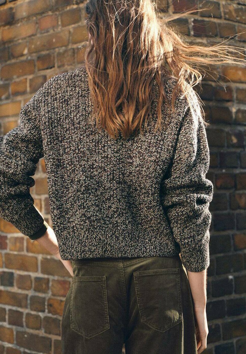 Brązowy sweter z fakturowanym splotem o kroju krótkim, połączony z oliwkowymi spodniami z kordurowej tkaniny, posiadającymi kieszenie z tyłu, na tle ceglanej ściany.