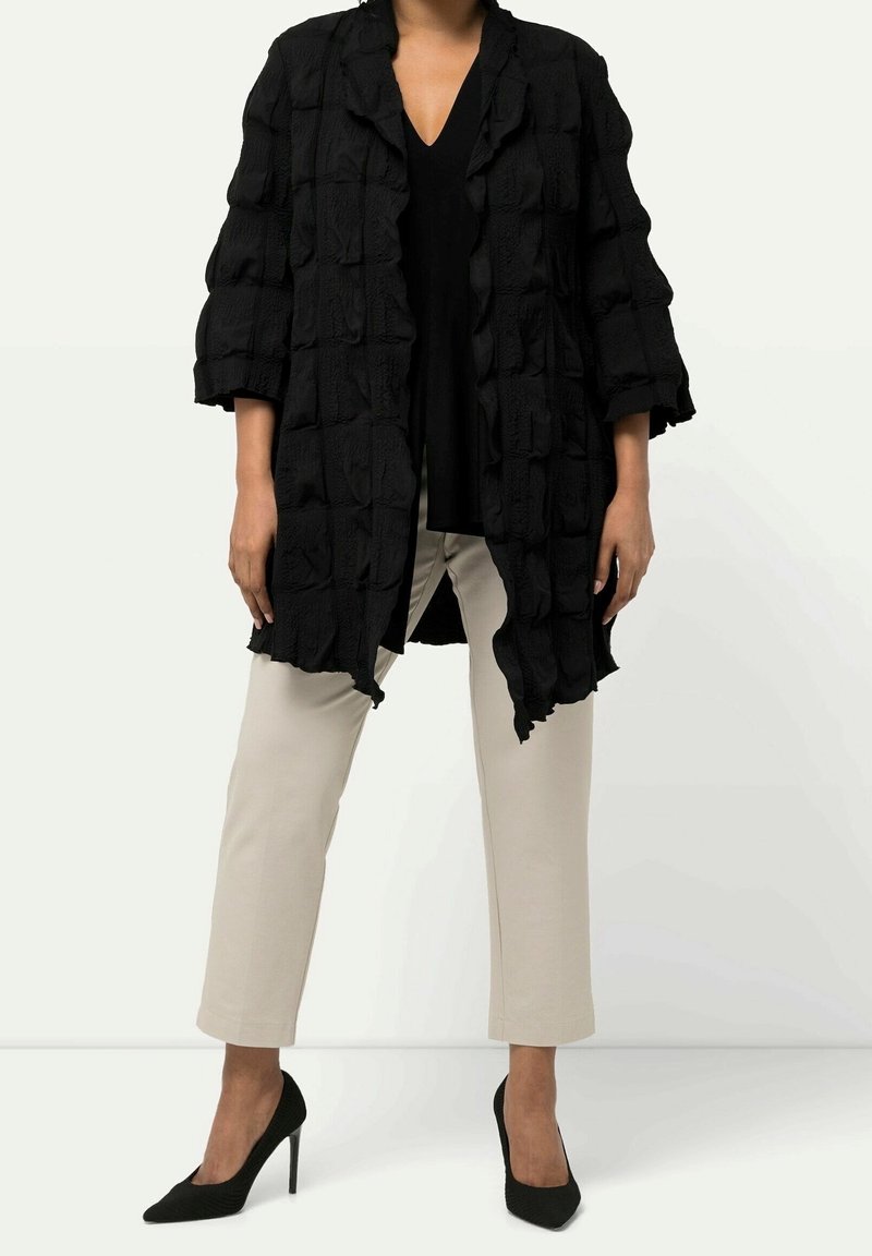 Black textured long cardigan with a loose fit, featuring a wavy design. Worn over a black top and beige pants, complemented by black heels.