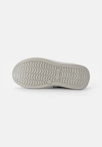 Beige Sneaker-Sohle mit einem strukturierten Zickzack-Muster. Verfügt über ein kreisförmiges Logo und minimale Markenkennzeichnung. Glatte Oberfläche mit leichten Grippdetails.