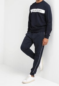 Námořnická mikina a tepláky vyrobené z bavlněného směsového materiálu, s bílým logem na pásce, žebrovanými manžetami a bočními detailem v barvě. V kombinaci s bílými teniskami.